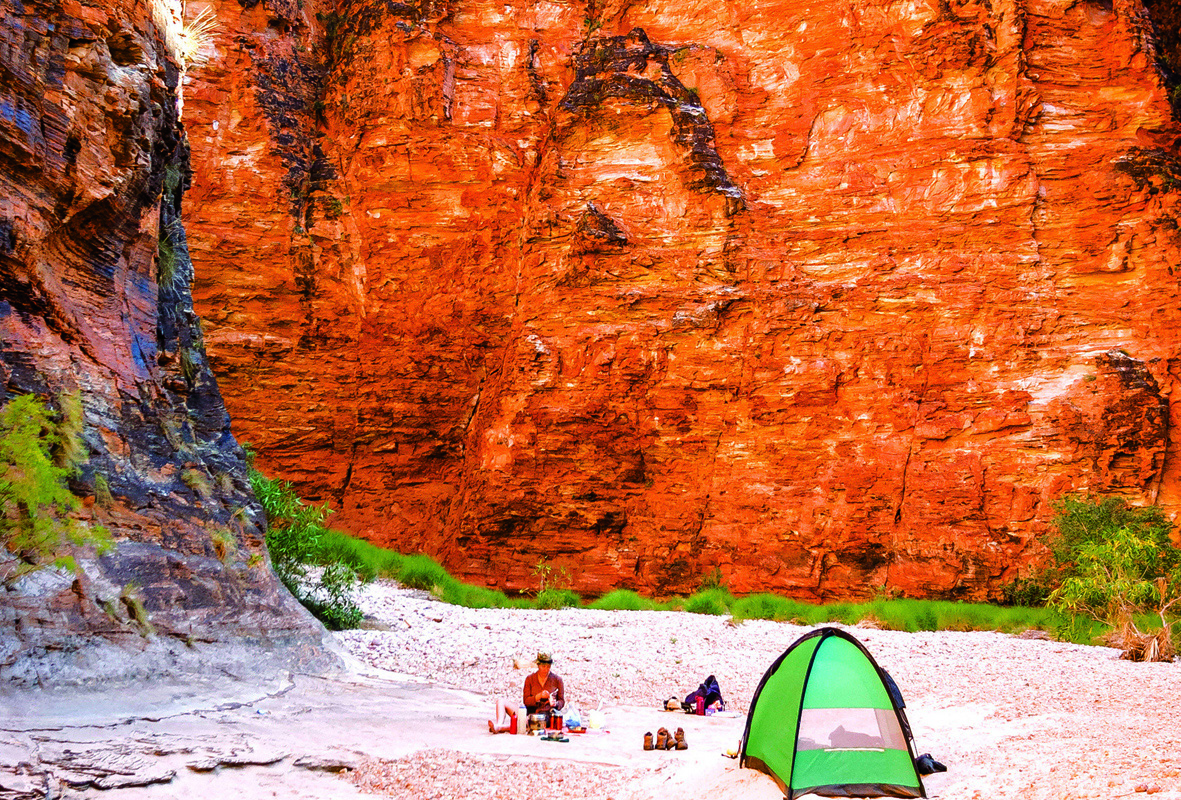 A person sits on the snad next to a green tent, at the base of a sandstone gorge